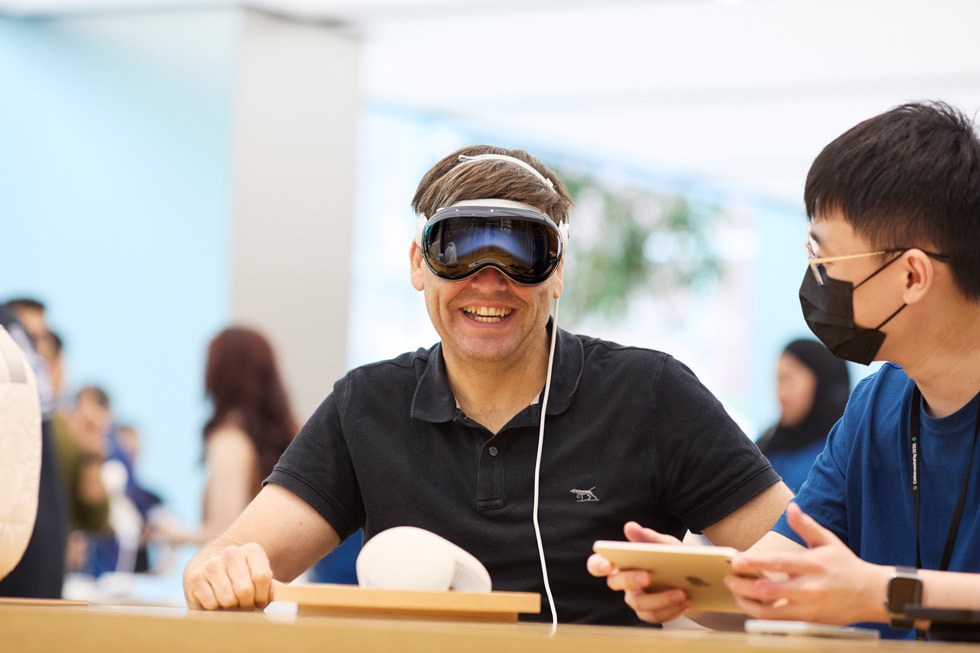 一位佩戴着 Apple Vision Pro 的顾客露出微笑，一名团队成员在一旁观看。