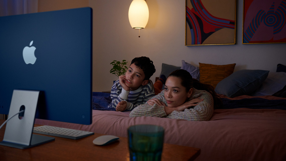 一名女性和一个孩子躺在床上，用新款 iMac 观看节目。