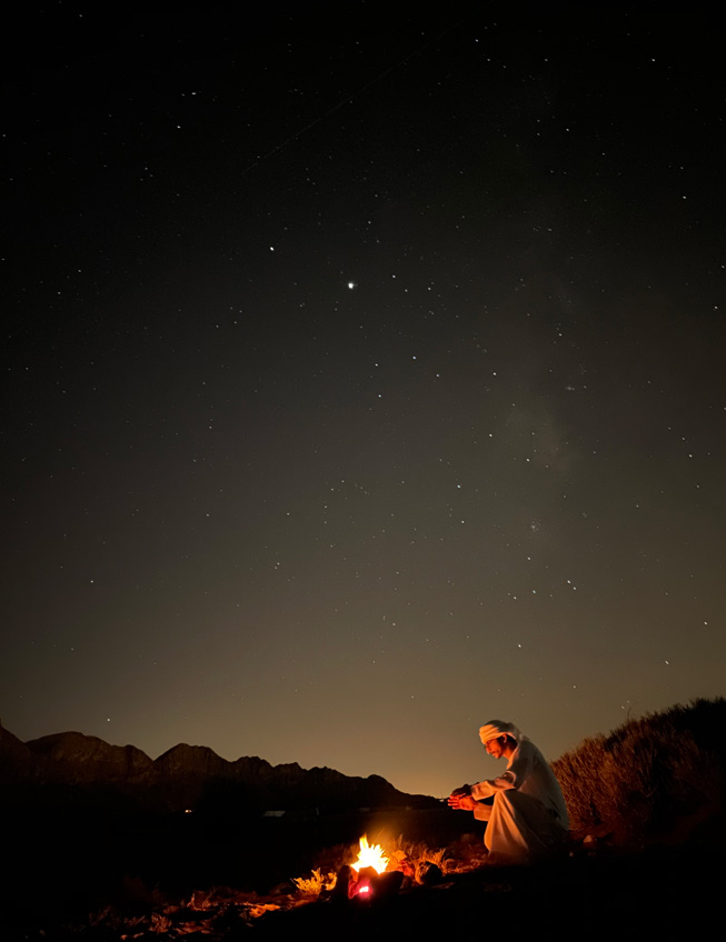一名男子坐在沙漠中营火前的夜间人像照。